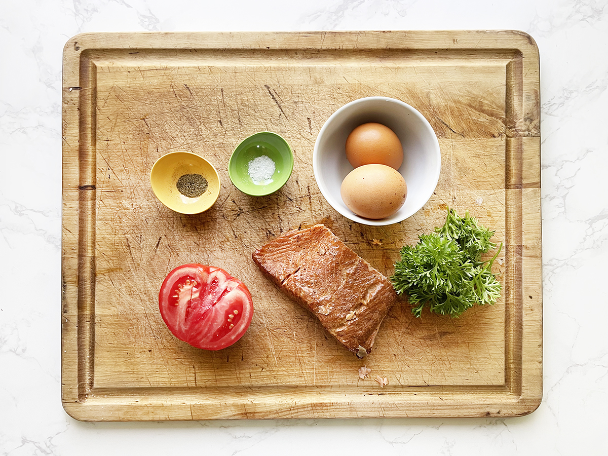 Cooked salmon, two eggs, a half of a tomato, parsley, salt and pepper on a cutting board.