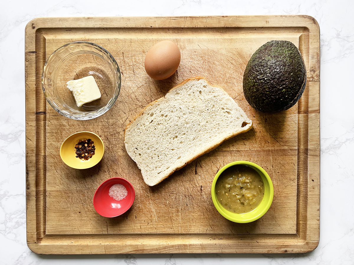 Cutting board with ingredients on top that will make a fried egg on toast with avocado and salsa.