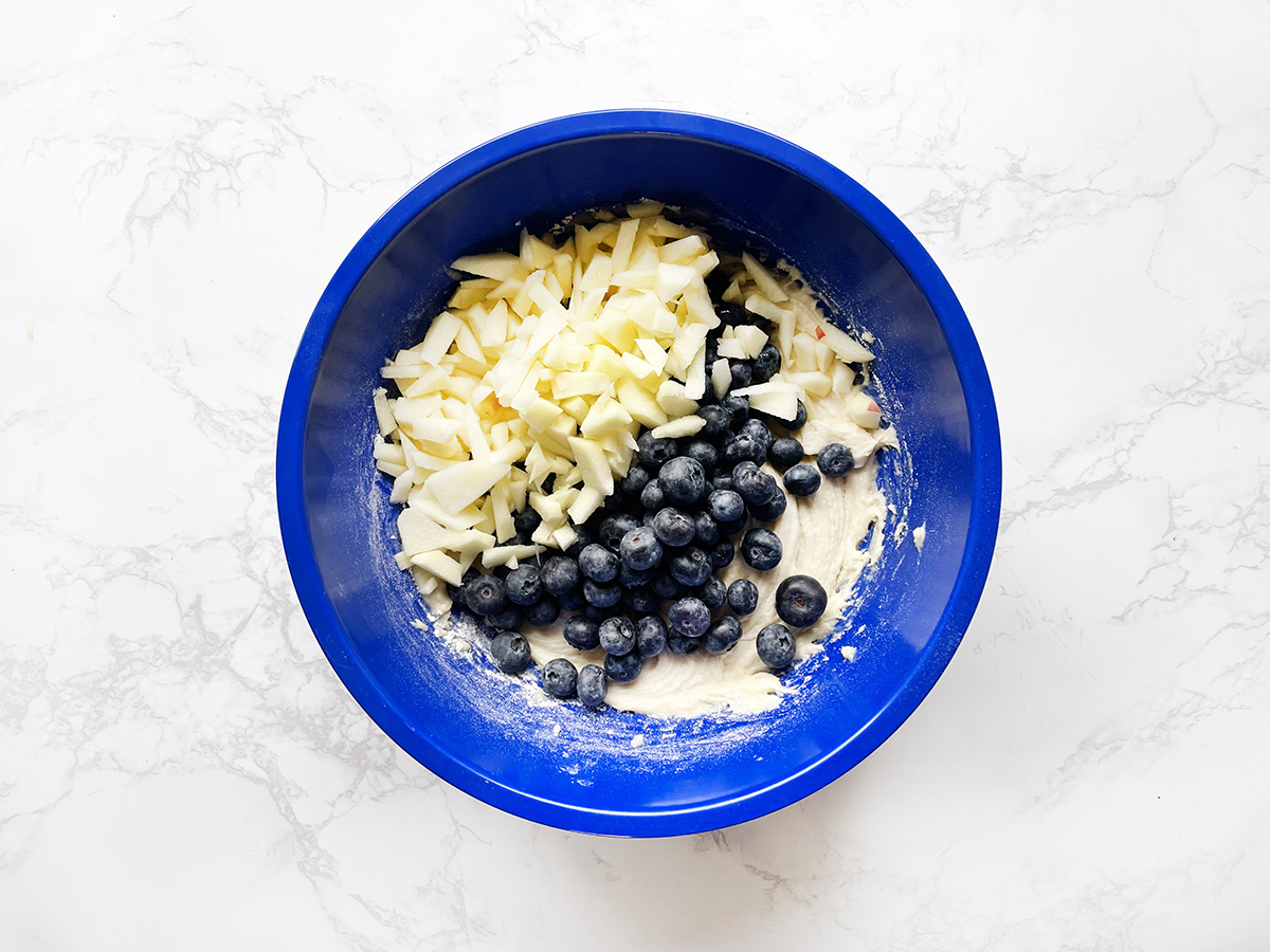 Muffin batter in a mixing bowl with blueberries and apples on top.