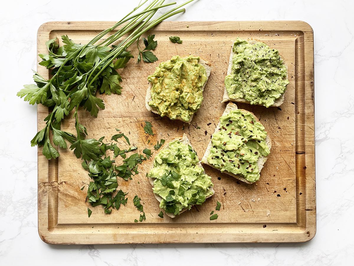 4 slices of avocado toast on a cutting board, all with different seasonings and sitting next to fresh parsley.