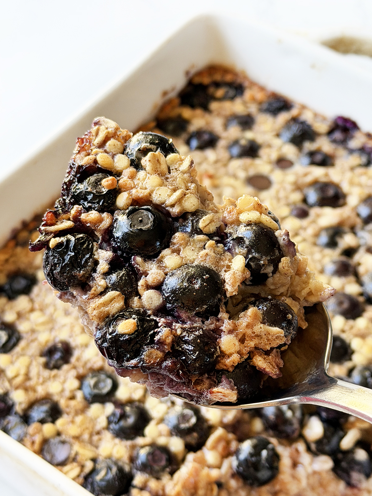 Spoonful of baked oatmeal with blueberries hovering over a baking pan.