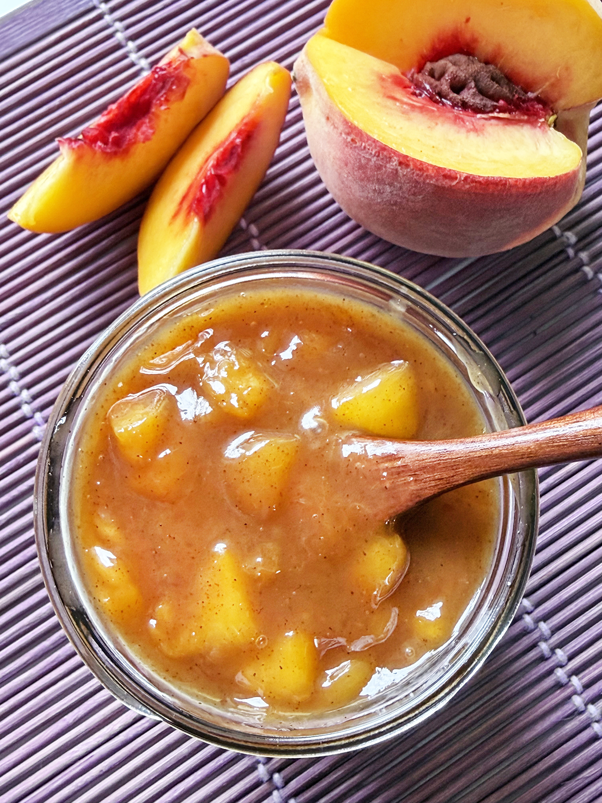 Mason jar of peach sauce with a spoon inside and peach nearby.