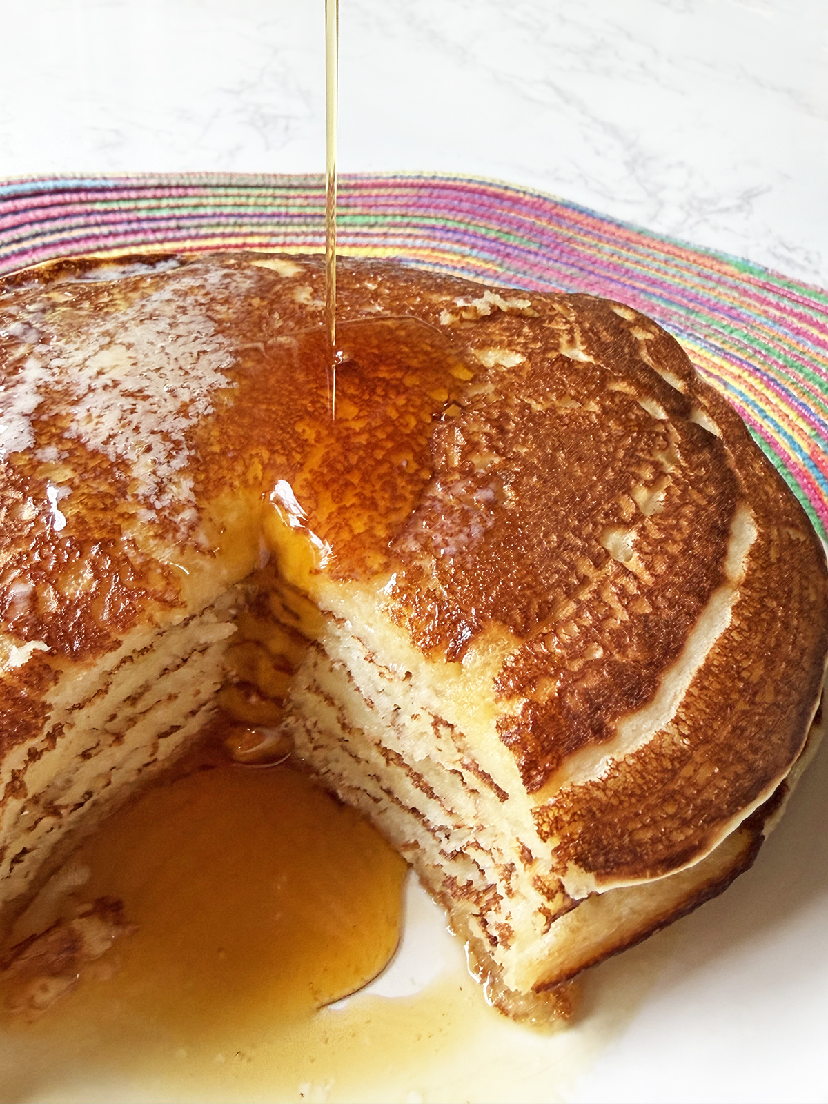 Syrup being poured onto a tall stack of pancakes that have a wedge removed.